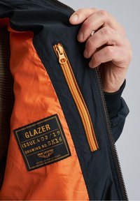 Marineblå og orange jakke indvendig med en lynlås lomme. Har et label med "GLAZER, ISSUE 402/19, DRAWING NO. SK16" og en tekstilstruktur.