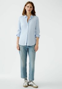 Light blue button-up shirt with long sleeves, worn with relaxed-fit faded jeans and grey socks, paired with white and black sneakers.