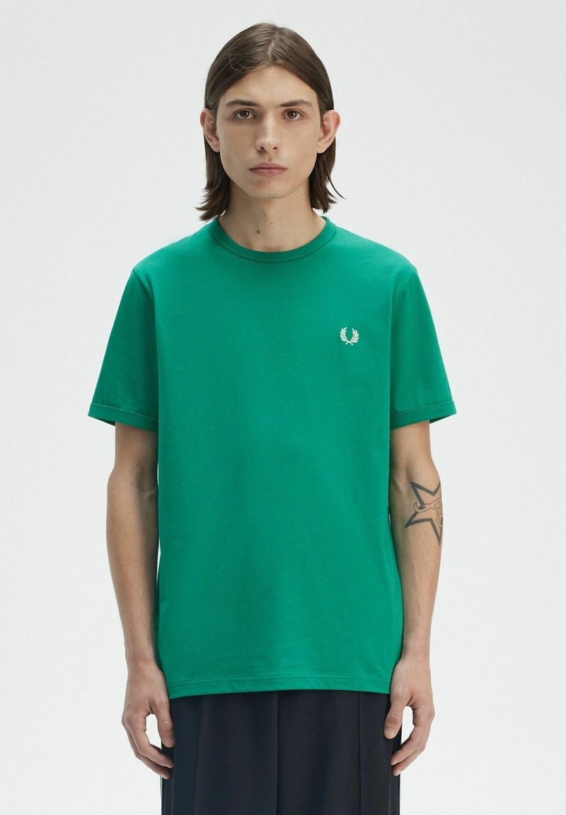 T-shirt verde in cotone con maniche corte, scollo rotondo e logo ricamato sul petto. Indossato con pantaloni neri fluidi.