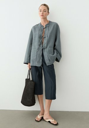 Femme portant une veste grise ample, pantalon court foncé, sandales beiges, tenant un grand sac cabas noir tissé, debout devant un mur blanc uni.