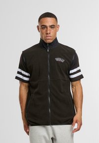 Gilet en polaire noir avec un col haut, une fermeture éclair à l'avant et des poches latérales ; présente des accents rayés blancs sur les manches courtes et un logo brodé.