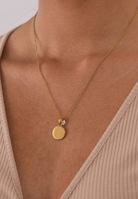 Goldkette mit einem runden Anhänger und einem kleinen quadratischen Akzent. Zarte Kette mit glänzender Oberfläche. Trägt man über einem strukturierten V-Ausschnitt-Oberteil.