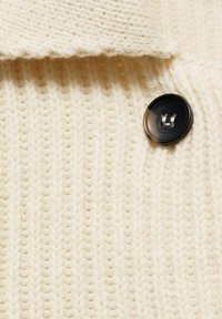 Tessuto lavorato a maglia beige con una trama a coste, caratterizzato da un bottone nero rotondo con due fori. Il lavoro a maglia ha un aspetto morbido e accogliente.