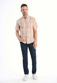 Korte mouwen geruite blouse in lichtoranje en blauwtonen, gecombineerd met donkerblauwe jeans en witte sneakers. Model staat met hand op de heup.