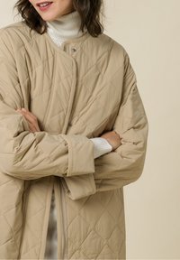 Abrigo acolchado beige con un diseño de rombos, con cierre de cremallera y mangas anchas. Se lleva sobre un suéter de cuello alto blanco y acanalado.