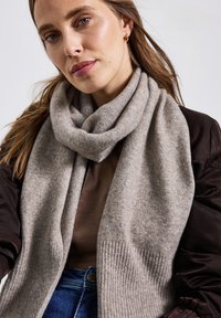 Kvinna med ljusbrunt hår som bär en tjock beige halsduk, brun jacka och blå jeans mot en neutral bakgrund.
