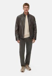 Bruine leren jas met een staande kraag, zijritszak en gestikte schouderdetails. Gedragen over een crèmekleurige trui en grijze broek.