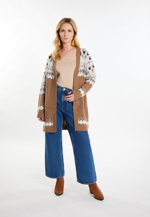 Langer Strickcardigan in Braun mit weißen geometrischen Mustern, kombiniert mit einem hellbeigen Oberteil und weit geschnittenen blauen Jeans, sowie braunen Stiefeletten.