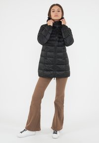 Giacca piumino nera con zip frontale, design trapuntato e cappuccio con pelliccia. Il modello indossa pantaloni a zampa d'elefante marroni e sneakers bianche.