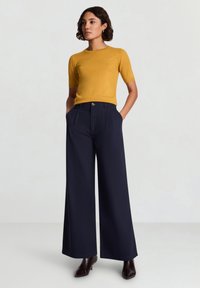 Senfgelbes kurzärmeliges Oberteil kombiniert mit hoch taillierter, weit geschnittener marineblauer Hose mit seitlichen Taschen und Knopfverschluss. Dunkle Pumps.