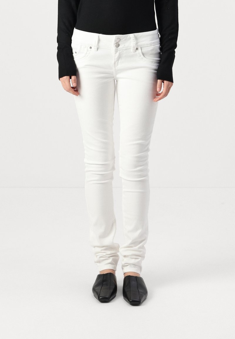 Jeans skinny blancs avec une texture lisse, design à cinq poches, quincaillerie argentée et fermeture à bouton standard, associés à des chaussures noires à enfiler.