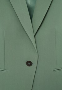 Veste en tissu vert à texture lisse, fermeture à un bouton foncé, et revers crantés. Comprend deux poches avant et une coupe ajustée.