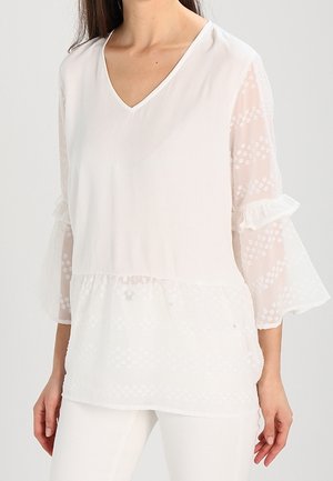 Femme portant un chemisier blanc avec des manches cloche transparentes et à motifs, et un pantalon blanc sur un fond uni.
