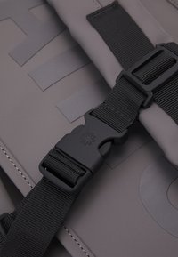 Gräudes Produkt aus Stoff mit einer schwarzen Schnalle und einem Riemen. Bemerkenswerte Details sind die Nähte und ein Logo auf dem Material, das eine glatte Textur aufweist.