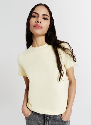 Giovane donna con lunghi capelli scuri che indossa una maglietta a maniche corte gialla chiara e jeans grigi, in piedi con le mani nelle tasche su uno sfondo bianco.