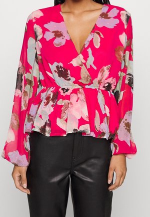 Nainen, joka pukeutunut kirkkaan pinkkiin, kukkakuvioiseen pitkähihnaiseen paitaan, jossa on V-aukko ja pepaum vyötärö, yhdistettynä mustiin nahkahousuihin.