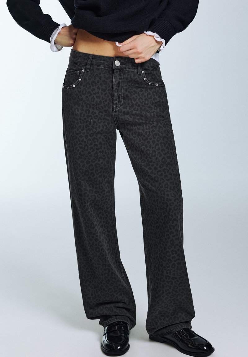 Personne portant un jean large imprimé léopard noir, des mocassins noirs brillants et un pull noir qu'elle soulève pour révéler le ventre.