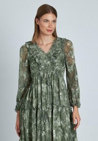 Vestido verde con estampado floral, con cuello y mangas con volantes. Presenta una falda en capas y una cintura ajustable con cordón. Fabricado en un tejido ligero y texturizado.