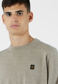 Maglione crew neck beige chiaro realizzato in morbido tessuto, con una piccola etichetta logo nera e oro sul lato sinistro.