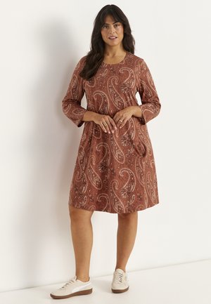 Frau trägt ein knielanges braunes Paisley-Kleid mit langen Ärmeln und weißen Sneakers, steht vor einem schlichten hellen Hintergrund.