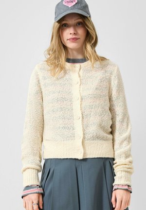 Giovane donna che indossa un cardigan leggero in maglia con bottoni, gonna grigia plissettata, camicia a righe con polsini e un berretto da baseball grigio con una toppa rosa.