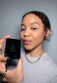 Czarna prostokątna kasetka z logo "dm" i napisem "Danessa Myricks Beauty". Modelka trzyma ją w jednej ręce, nosząc złotą biżuterię.