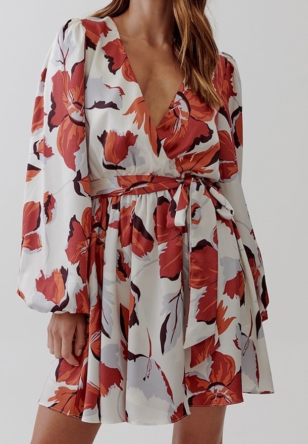Femme portant une robe blanche avec de grands motifs floraux rouges et orange, des manches longues bouffantes et une ceinture nouée à la taille, debout devant un fond uni.
