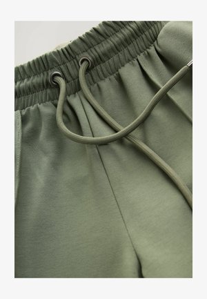 Tamsiai žali sportiniai šortai su elastingu juosmeniu, kuris turi raištį su metaliniais galiukais ir minkšta tekstūra.