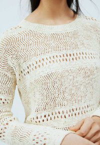 Pull tricoté beige clair présentant un motif texturé avec un tissage ajouré et un design en zigzag, manches longues et encolure ronde.