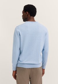 Mann, der mit dem Rücken zur Kamera steht, trägt einen hellblauen Langarmpullover und braune Hosen vor einem schlichten beigen Hintergrund.