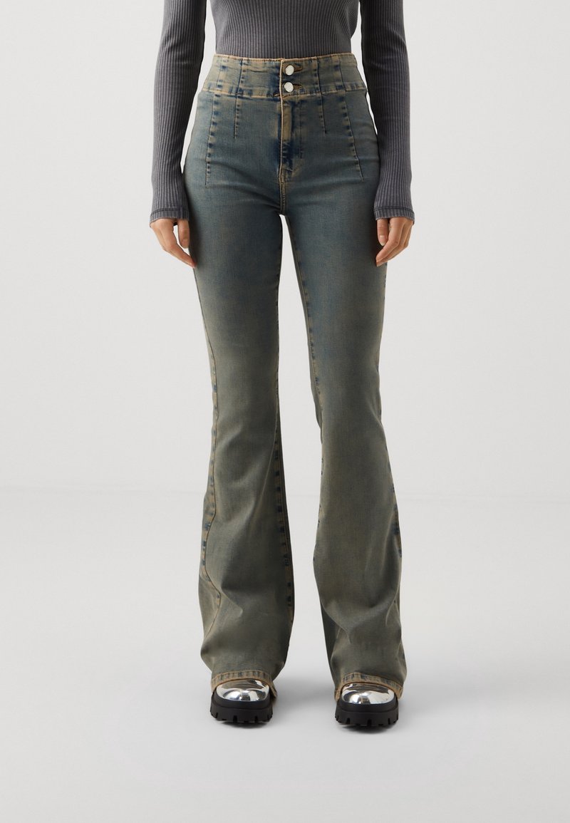 Jean évasé vert délavé taille haute porté avec un top gris côtelé à manches longues et des bottes noires épaisses avec embouts métalliques.