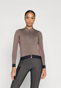 Giacca da ciclismo marrone con dettagli rosa, zip intera, maniche lunghe e polsini neri. Abbinata a pantaloni da ciclismo imbottiti marroni scuri.