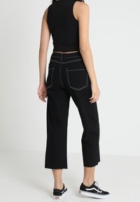 Czarne, krótkie dżinsy z widocznym szwem, luźny krój i postrzępione końce. Połączone z czarnymi trampkami, widziane z tyłu na jednolitym tle.