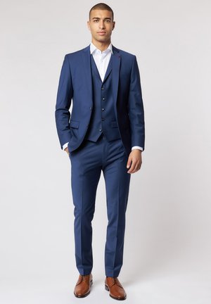 Navy blue three-piece suit, slim fit, composed of fabric with a subtle texture. Brown leather shoes complement the outfit. White dress shirt visible.