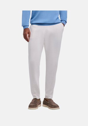 Mann trägt hellgraue schmale Hose, blauen Pullover und braune Wildleder-Slipper und steht mit einer Hand in der Tasche.
