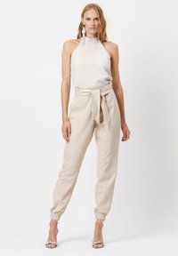 Halterneck top van lichtcrème stof, gecombineerd met beige tapered broek met een strikceintuur en elastische manchetten. Geaccentueerd met zilveren oorbellen.
