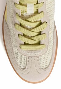 Zapatilla ligera con parte superior de tejido beige, acentos amarillos y cordones verdes. Detalles de ante suave y suela de goma texturizada.