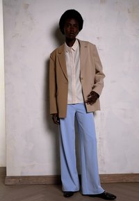 Beige übergroßer Blazer über einem hellen Hemd, kombiniert mit weit geschnittenen hellblauen Hosen. Schwarze Slipper, vor einem neutralen Hintergrund.