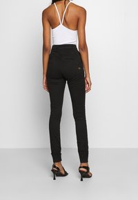 Jeans ajustados negros de cintura alta con un acabado texturizado, que cuentan con un bolsillo trasero y puños enrollados, combinados con una blusa blanca de tirantes finos.