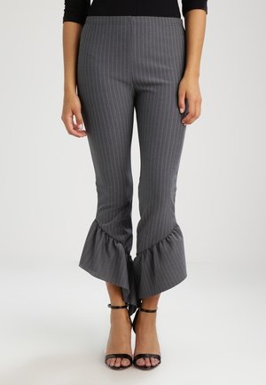 Kobieta ubrana w szare spodnie w prążki z falbankami na rozszerzanych nogawkach oraz czarne szpilki z paskiem na kostkę, stojąca na tle jednolitego jasnego tła.