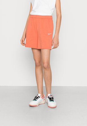 Osoba w pomarańczowych szortach Nike z białym logo oraz białych butach sportowych z różowymi i czarnymi detalami, stojąca na jednolitym tle.