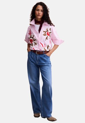 Mujer con cabello rizado que lleva una camisa rosa con estampado floral, bufanda granate, vaqueros azules de pierna ancha, cinturón marrón y zapatos marrones sobre fondo blanco.
