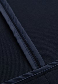 Gros plan d'un vêtement en tissu foncé avec une fermeture éclair centrale et des coutures surpiquées le long des bords.