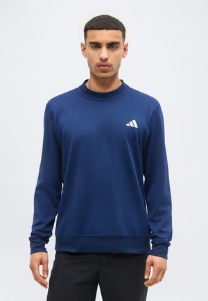 Muž v námorníckom modrom pulóvri s dlhým rukávom a bielym trojuholníkovým logom na hrudi stojaci pred jednoduchým svetlosivým pozadím.