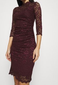 Robe en dentelle bordeaux avec des manches trois-quarts, corsage ajusté, ourlet festonné et détail froncé sur le côté, présentant un motif floral.