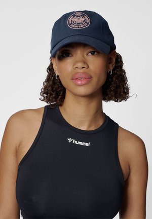 Młoda kobieta o kręconych włosach nosi granatową czapkę z logo pszczoły oraz czarny sportowy top bez rękawów z logo „hummel” na prostym tle.