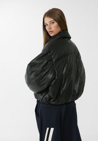 Giacca di pelle nera oversize con colletto alto, texture trapuntata e maniche cadute. Abbinata a pantaloni blu navy con strisce bianche.