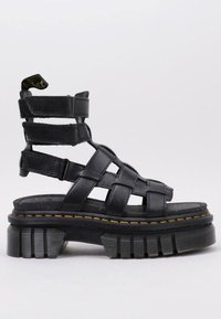 Sandalia negra estilo gladiador con suela gruesa y estriada, tres correas de Velcro ajustables y un tirador en el tobillo con logotipo amarillo.