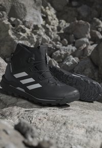Sorte Adidas Terrex vandrestøvler med hvide striber placeret på klippefyldt terræn, der viser robust sål og ankelhøjt design.
