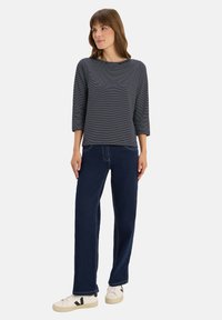 Femme debout portant un haut à rayures noir et blanc à manches longues, un jean bleu foncé et des baskets blanches avec des logos "V" noirs.